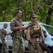 A group of military or tactical personnel in various camouflage uniforms are gathered in a wooded outdoor area near a white van. Two central figures appear to be reviewing a document or map together, suggesting a briefing or planning session. The personnel are equipped with tactical vests, pouches, and sidearms, indicating an operational or training exercise.