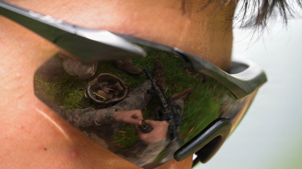 A close-up photograph of a person wearing olive/green-framed tactical sunglasses, with a detailed reflection visible in the lens. The reflection shows a military personnel in camouflage uniform handling a rifle and ammunition on a grassy area, with a helmet and gear visible on the ground. The creative composition uses the sunglass lens as a mirror to capture the scene.