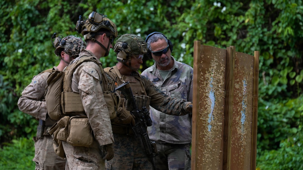 A group of military personnel in full combat gear are gathered at a shooting range, examining a bullet-riddled wooden target board. An instructor or range officer in civilian-style camouflage clothing and hearing protection appears to be providing feedback or instruction. The setting appears to be an outdoor range surrounded by dense tropical vegetation.