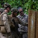 A group of military personnel in full combat gear are gathered at a shooting range, examining a bullet-riddled wooden target board. An instructor or range officer in civilian-style camouflage clothing and hearing protection appears to be providing feedback or instruction. The setting appears to be an outdoor range surrounded by dense tropical vegetation.