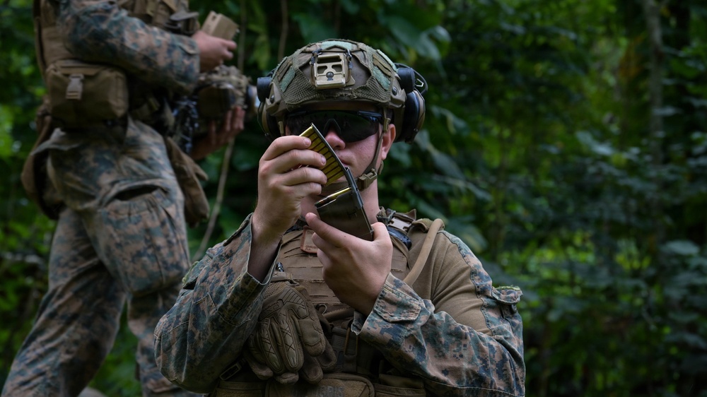 A U.S. Marine in full combat gear is inspecting or loading a rifle magazine with visible brass cartridges in a jungle or forested environment. A second Marine in matching MARPAT camouflage uniform is visible in the background. The setting appears to be a field training exercise or operational patrol in a tropical location.