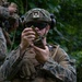 A U.S. Marine in full combat gear is inspecting or loading a rifle magazine with visible brass cartridges in a jungle or forested environment. A second Marine in matching MARPAT camouflage uniform is visible in the background. The setting appears to be a field training exercise or operational patrol in a tropical location.