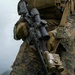 A military service member in MARPAT (Marine pattern) digital camouflage uniform is holding a modern assault rifle with multiple tactical accessories. The image is a close-up torso shot focusing on the weapon and gear, with a overcast sky and vegetation visible in the background. The equipment and uniform pattern are consistent with U.S. Marine Corps kit.