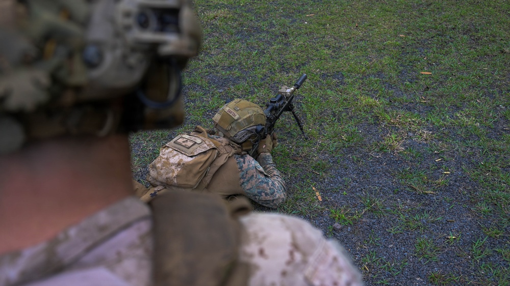 A military service member in MARPAT desert camouflage is in a prone firing position on a grassy/gravel surface, aiming a rifle equipped with a bipod. The image is taken from the perspective of a second service member in the foreground wearing a helmet with night vision device mount. This appears to be a training exercise or range activity.