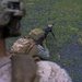 A military service member in MARPAT desert camouflage is in a prone firing position on a grassy/gravel surface, aiming a rifle equipped with a bipod. The image is taken from the perspective of a second service member in the foreground wearing a helmet with night vision device mount. This appears to be a training exercise or range activity.