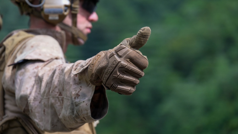 A military personnel wearing desert camouflage (MARPAT pattern) uniform is giving a thumbs-up gesture toward the camera. The individual is equipped with tactical gear including a helmet with eye protection and a tactical vest. The background consists of blurred green foliage, suggesting an outdoor training or operational environment.