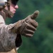 A military personnel wearing desert camouflage (MARPAT pattern) uniform is giving a thumbs-up gesture toward the camera. The individual is equipped with tactical gear including a helmet with eye protection and a tactical vest. The background consists of blurred green foliage, suggesting an outdoor training or operational environment.