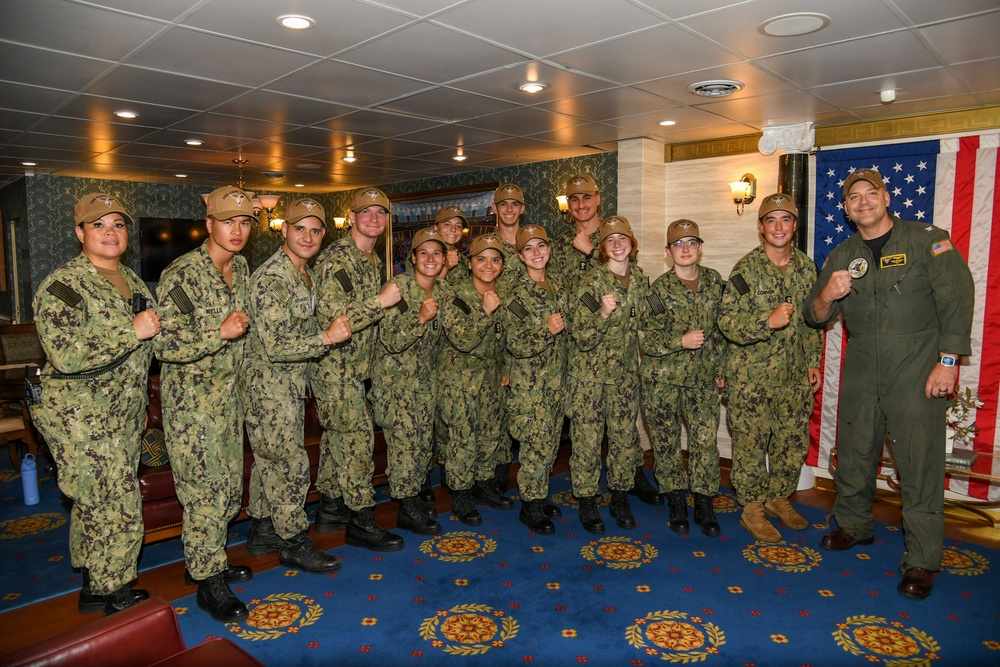 A group of approximately 14 U.S. Navy personnel pose for a group photo in what appears to be an interior room aboard a naval vessel or military facility. Most are wearing Navy Working Uniform (NWU Type III) camouflage with tan covers, while one individual on the far right wears an olive green flight suit. The group is posing with raised fists in a celebratory or solidarity gesture in front of an American flag.