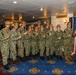 A group of approximately 14 U.S. Navy personnel pose for a group photo in what appears to be an interior room aboard a naval vessel or military facility. Most are wearing Navy Working Uniform (NWU Type III) camouflage with tan covers, while one individual on the far right wears an olive green flight suit. The group is posing with raised fists in a celebratory or solidarity gesture in front of an American flag.