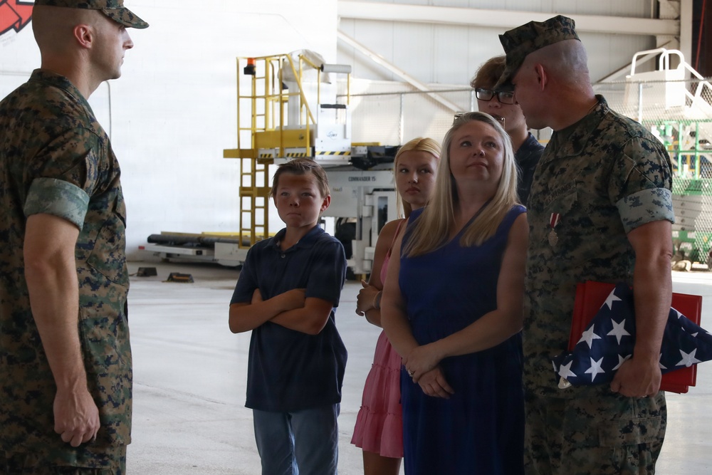 Two U.S. Marines in camouflage uniforms are interacting with a group of civilians, including children and a woman, inside what appears to be a military hangar. One Marine on the right is holding a folded American flag, suggesting a formal military ceremony such as a retirement, promotion, or commendation event. The civilians appear to be family members attending the ceremony.