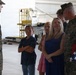 Two U.S. Marines in camouflage uniforms are interacting with a group of civilians, including children and a woman, inside what appears to be a military hangar. One Marine on the right is holding a folded American flag, suggesting a formal military ceremony such as a retirement, promotion, or commendation event. The civilians appear to be family members attending the ceremony.