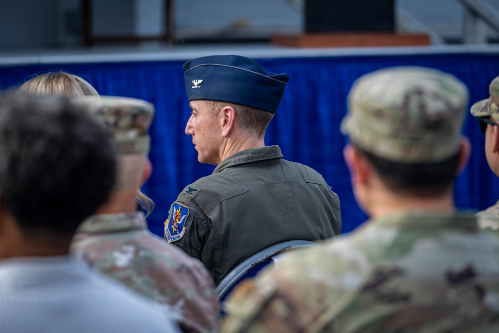 The image shows a group of military personnel at what appears to be a formal military event or ceremony. The central figure is a U.S. Air Force Colonel, identifiable by the silver eagle insignia on his flight cap and a unit patch on his flight suit. Surrounding personnel are wearing Army Combat Uniforms (ACU/OCP pattern), suggesting a joint-service gathering.