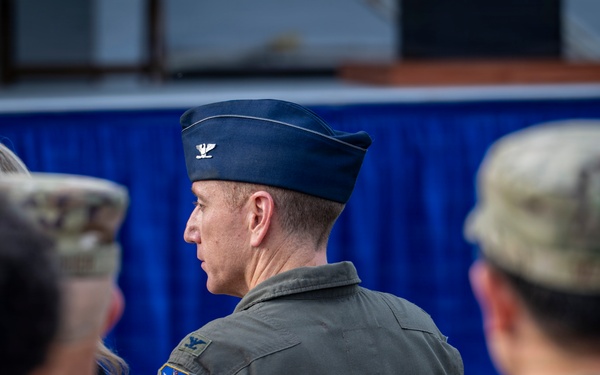 The image shows a group of military personnel at what appears to be a formal military event or ceremony. The central figure is a U.S. Air Force Colonel, identifiable by the silver eagle insignia on his flight cap and a unit patch on his flight suit. Surrounding personnel are wearing Army Combat Uniforms (ACU/OCP pattern), suggesting a joint-service gathering.