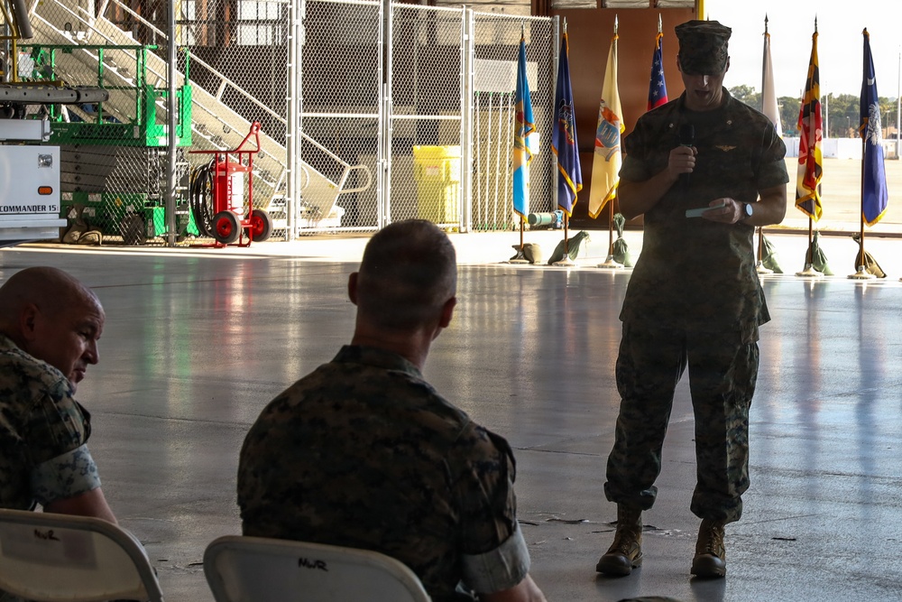 A military ceremony taking place inside what appears to be an aircraft hangar, with a standing Marine officer addressing seated personnel. Multiple ceremonial flags are displayed in the background, suggesting a formal military event such as a change of command or award ceremony. Three Marines in camouflage uniforms are visible, with one standing and speaking while two others are seated.