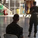 A military ceremony taking place inside what appears to be an aircraft hangar, with a standing Marine officer addressing seated personnel. Multiple ceremonial flags are displayed in the background, suggesting a formal military event such as a change of command or award ceremony. Three Marines in camouflage uniforms are visible, with one standing and speaking while two others are seated.