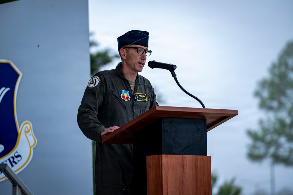 A U.S. Air Force officer in a flight suit is speaking at a podium with a microphone during what appears to be an official military ceremony or event. He is wearing a service cap, glasses, and multiple patches on his olive-green flight suit. A partial military unit shield/emblem is visible on a backdrop to his left.