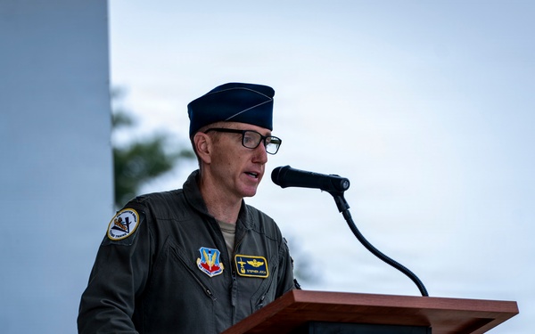 A U.S. Air Force officer in a flight suit is speaking at a podium with a microphone during what appears to be an official military ceremony or event. He is wearing a service cap, glasses, and multiple patches on his olive-green flight suit. A partial military unit shield/emblem is visible on a backdrop to his left.