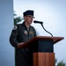 A U.S. Air Force officer in a flight suit is speaking at a podium with a microphone during what appears to be an official military ceremony or event. He is wearing a service cap, glasses, and multiple patches on his olive-green flight suit. A partial military unit shield/emblem is visible on a backdrop to his left.