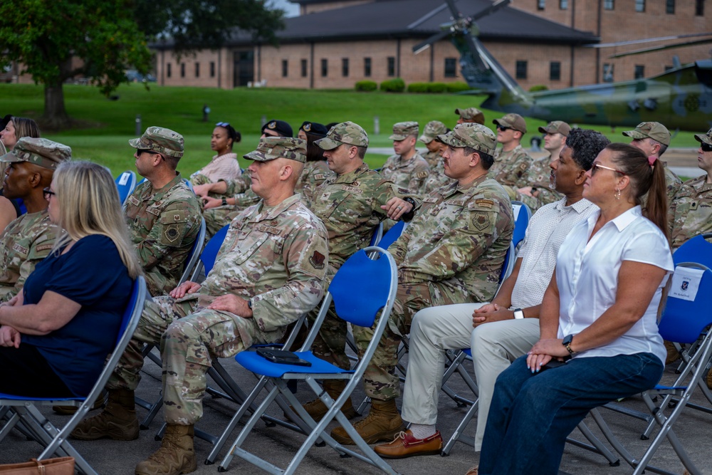 A group of military personnel in OCP (Operational Camouflage Pattern) uniforms and civilians are seated in blue folding chairs outdoors, appearing to attend a formal military ceremony or event. The setting appears to be on a military installation, with a brick building and a static display helicopter visible in the background. The audience is attentively facing toward an unseen speaker or stage.