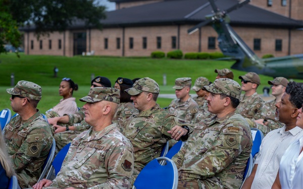 A group of military personnel in OCP (Operational Camouflage Pattern) uniforms and civilians are seated in blue folding chairs outdoors, appearing to attend a formal military ceremony or event. The setting appears to be on a military installation, with a brick building and a static display helicopter visible in the background. The audience is attentively facing toward an unseen speaker or stage.