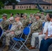 A group of military personnel in OCP (Operational Camouflage Pattern) uniforms and civilians are seated in blue folding chairs outdoors, appearing to attend a formal military ceremony or event. The setting appears to be on a military installation, with a brick building and a static display helicopter visible in the background. The audience is attentively facing toward an unseen speaker or stage.