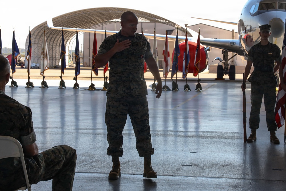A military ceremony is taking place inside or at the entrance of an aircraft hangar on a flight line. A Marine in camouflage utilities stands at the center, appearing to address an audience while placing his hand on his chest. Multiple ceremonial flags are displayed in the background alongside a parked aircraft.