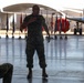 A military ceremony is taking place inside or at the entrance of an aircraft hangar on a flight line. A Marine in camouflage utilities stands at the center, appearing to address an audience while placing his hand on his chest. Multiple ceremonial flags are displayed in the background alongside a parked aircraft.