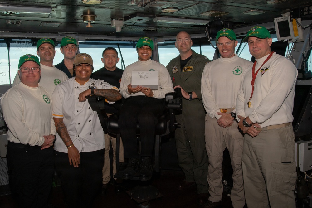 A group of approximately nine individuals pose for a group photo on what appears to be the bridge of a naval vessel. Most are wearing matching white long-sleeve shirts with green 'SAFETY' caps, while one individual wears a chef's coat and another wears an olive green flight suit. A person seated in the center holds a white card with handwritten text.
