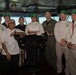 A group of approximately nine individuals pose for a group photo on what appears to be the bridge of a naval vessel. Most are wearing matching white long-sleeve shirts with green 'SAFETY' caps, while one individual wears a chef's coat and another wears an olive green flight suit. A person seated in the center holds a white card with handwritten text.
