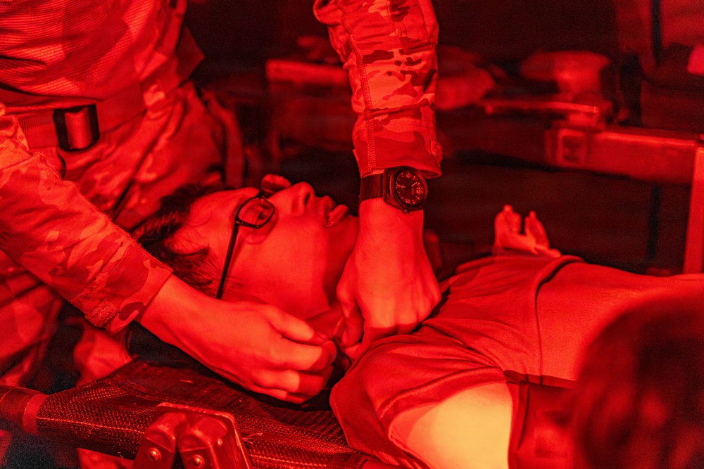 The image shows a military medical scenario illuminated by red lighting, where a person in camouflage uniform is attending to a casualty lying on what appears to be a litter or stretcher. The attendant is performing what appears to be a medical assessment or treatment on the supine individual. The red lighting is consistent with night operations or aircraft interior lighting used to preserve night vision.