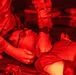 The image shows a military medical scenario illuminated by red lighting, where a person in camouflage uniform is attending to a casualty lying on what appears to be a litter or stretcher. The attendant is performing what appears to be a medical assessment or treatment on the supine individual. The red lighting is consistent with night operations or aircraft interior lighting used to preserve night vision.