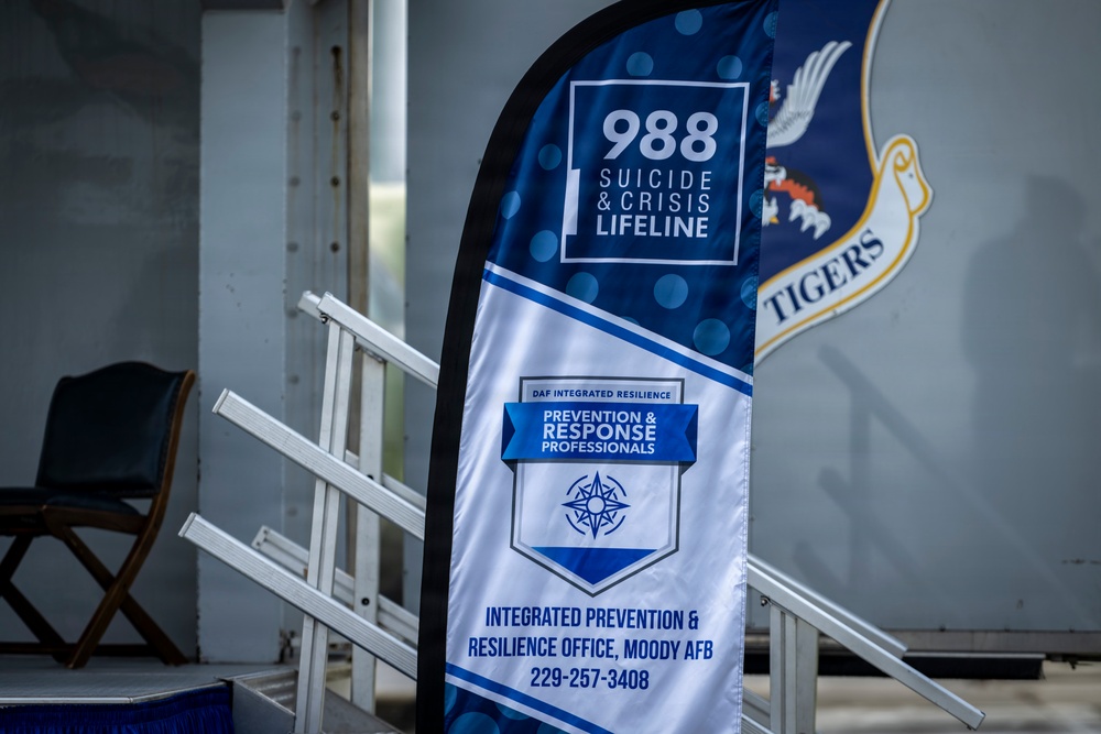 The image shows a promotional banner at Moody Air Force Base promoting the 988 Suicide &amp; Crisis Lifeline alongside the DAF Integrated Resilience Prevention &amp; Response Professionals program. The banner is displayed near a ramp or bleacher structure, with a partial Air Force shield emblem visible in the background bearing the word 'TIGERS'. The setting appears to be an indoor or covered military facility.