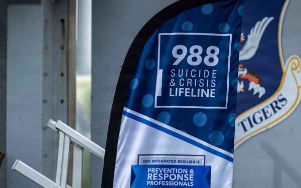 The image shows a promotional banner at Moody Air Force Base promoting the 988 Suicide &amp; Crisis Lifeline alongside the DAF Integrated Resilience Prevention &amp; Response Professionals program. The banner is displayed near a ramp or bleacher structure, with a partial Air Force shield emblem visible in the background bearing the word 'TIGERS'. The setting appears to be an indoor or covered military facility.