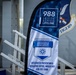 The image shows a promotional banner at Moody Air Force Base promoting the 988 Suicide &amp; Crisis Lifeline alongside the DAF Integrated Resilience Prevention &amp; Response Professionals program. The banner is displayed near a ramp or bleacher structure, with a partial Air Force shield emblem visible in the background bearing the word 'TIGERS'. The setting appears to be an indoor or covered military facility.