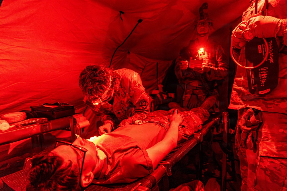 Military medical personnel are treating a casualty on a field stretcher inside a red-lit tactical medical tent. At least three personnel in camouflage uniforms are attending to the patient, with one leaning over performing a procedure, one holding an IV bag, and one providing illumination with a headlamp. The red lighting is consistent with tactical/blackout medical operations to preserve night vision.