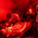 Military medical personnel are treating a casualty on a field stretcher inside a red-lit tactical medical tent. At least three personnel in camouflage uniforms are attending to the patient, with one leaning over performing a procedure, one holding an IV bag, and one providing illumination with a headlamp. The red lighting is consistent with tactical/blackout medical operations to preserve night vision.