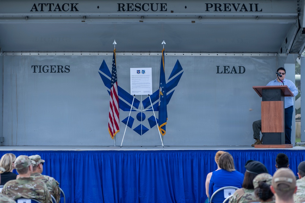 A formal military ceremony is taking place on an outdoor stage at what appears to be a U.S. Air Force installation. A speaker stands at a wooden podium addressing an audience of military personnel and civilians seated in front of the stage. The stage backdrop displays the U.S. Air Force symbol along with the motto 'Attack · Rescue · Prevail' and the phrase 'Tigers Lead'.