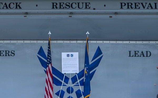A formal military ceremony is taking place on an outdoor stage at what appears to be a U.S. Air Force installation. A speaker stands at a wooden podium addressing an audience of military personnel and civilians seated in front of the stage. The stage backdrop displays the U.S. Air Force symbol along with the motto 'Attack · Rescue · Prevail' and the phrase 'Tigers Lead'.