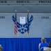 A formal military ceremony is taking place on an outdoor stage at what appears to be a U.S. Air Force installation. A speaker stands at a wooden podium addressing an audience of military personnel and civilians seated in front of the stage. The stage backdrop displays the U.S. Air Force symbol along with the motto 'Attack · Rescue · Prevail' and the phrase 'Tigers Lead'.