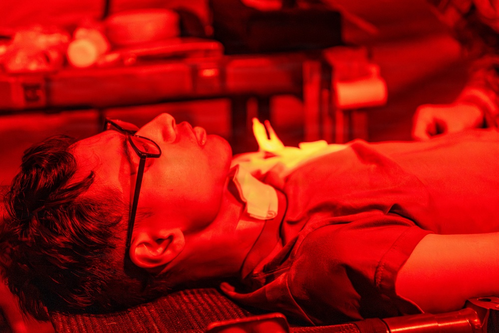 A person is lying on their back under intense red lighting, wearing glasses and what appears to be eye protection patches on their face. The scene suggests a medical, therapeutic, or cosmetic treatment setting, possibly red light therapy or a similar procedure. Another person's hand is partially visible in the background, suggesting an attendant or practitioner is present.