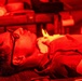 A person is lying on their back under intense red lighting, wearing glasses and what appears to be eye protection patches on their face. The scene suggests a medical, therapeutic, or cosmetic treatment setting, possibly red light therapy or a similar procedure. Another person's hand is partially visible in the background, suggesting an attendant or practitioner is present.