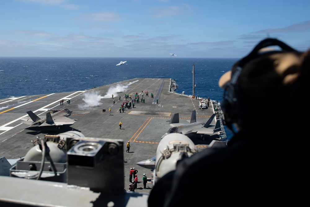An aerial view from an elevated position on a naval aircraft carrier shows active flight deck operations in open ocean. Multiple F-35 aircraft are visible on the deck and in flight, with deck crew in color-coded jerseys managing launch operations. Steam or smoke from a catapult launch is visible mid-deck, with at least two aircraft already airborne in the background.