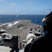 An aerial view from an elevated position on a naval aircraft carrier shows active flight deck operations in open ocean. Multiple F-35 aircraft are visible on the deck and in flight, with deck crew in color-coded jerseys managing launch operations. Steam or smoke from a catapult launch is visible mid-deck, with at least two aircraft already airborne in the background.