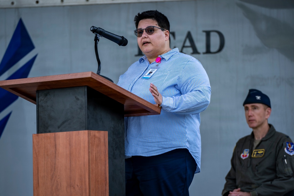 A civilian speaker addresses an audience from a wooden podium equipped with a microphone at what appears to be a military installation event. The speaker wears a blue striped shirt, sunglasses, and a visible ID badge with a pink accent. A uniformed military officer in olive flight suit and service cap is visible in the background to the right.