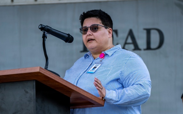 A civilian speaker addresses an audience from a wooden podium equipped with a microphone at what appears to be a military installation event. The speaker wears a blue striped shirt, sunglasses, and a visible ID badge with a pink accent. A uniformed military officer in olive flight suit and service cap is visible in the background to the right.