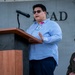 A civilian speaker addresses an audience from a wooden podium equipped with a microphone at what appears to be a military installation event. The speaker wears a blue striped shirt, sunglasses, and a visible ID badge with a pink accent. A uniformed military officer in olive flight suit and service cap is visible in the background to the right.