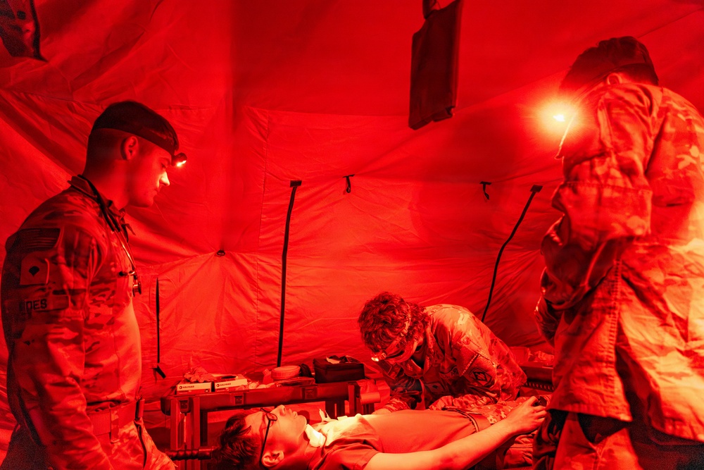 Military personnel are conducting a medical treatment procedure inside a field tent illuminated entirely by red light. Three soldiers in camouflage uniforms, equipped with headlamps, are attending to a patient lying on a field stretcher or cot. The red lighting environment is consistent with tactical night operations medical training or field triage.