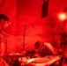 Military personnel are conducting a medical treatment procedure inside a field tent illuminated entirely by red light. Three soldiers in camouflage uniforms, equipped with headlamps, are attending to a patient lying on a field stretcher or cot. The red lighting environment is consistent with tactical night operations medical training or field triage.