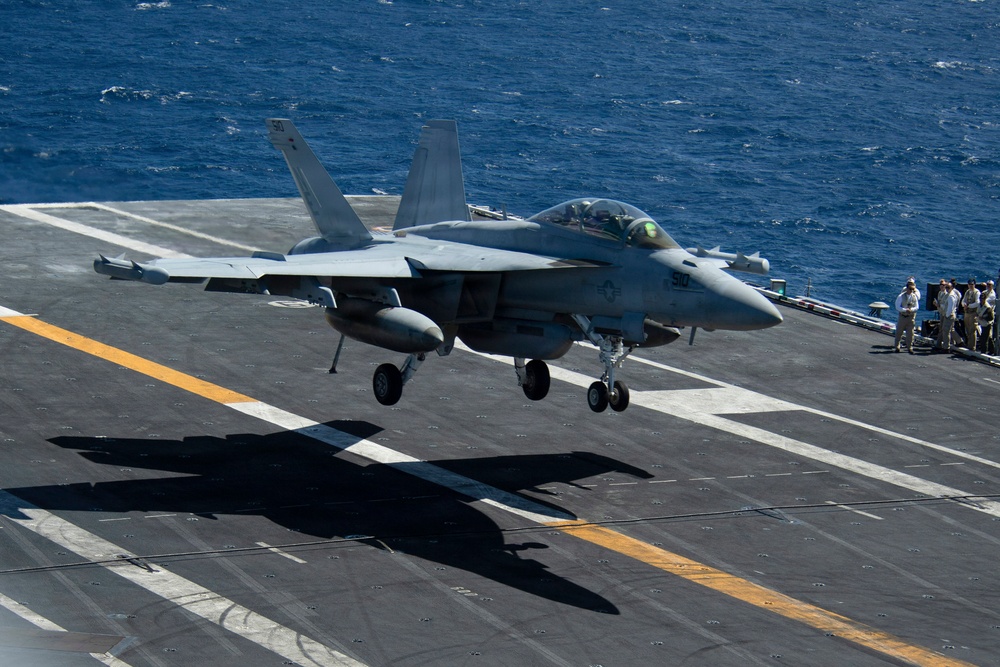 A U.S. Navy carrier-based jet aircraft, consistent with an F/A-18 Super Hornet, is captured moments before landing on an aircraft carrier flight deck. The aircraft's landing gear is fully deployed and it appears to be in the final approach phase over the carrier deck. Several flight deck personnel are visible on the starboard side observing the operation.