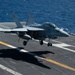 A U.S. Navy carrier-based jet aircraft, consistent with an F/A-18 Super Hornet, is captured moments before landing on an aircraft carrier flight deck. The aircraft's landing gear is fully deployed and it appears to be in the final approach phase over the carrier deck. Several flight deck personnel are visible on the starboard side observing the operation.